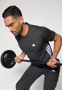 Camiseta deportiva negra con detalles de tres rayas blancas, sosteniendo una barra con un disco de peso, vistiendo pantalones grises, postura concentrada.