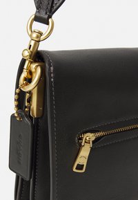 Sac à main en cuir noir avec quincaillerie dorée, comportant une poche zippée, un porte-clés en métal et une étiquette portant la marque "Coach". Texture lisse, design structuré.