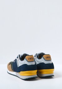 Baskets avec des matériaux mélangés : daim bleu marine et marron, tissu gris, et un onglet de talon beige avec "1973" imprimé. Semelle extérieure jaune, bande de roulement en caoutchouc texturé.