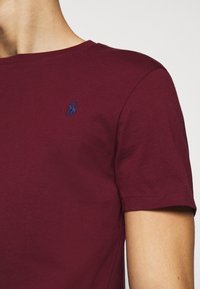 Burgundy puuvillane t-särk, millel on ümmargune kaelus. Esiküljel on väike meresinine brodeeritud logo vasakul rinnal ja lühikesed varrukad. Sile tekstuur.