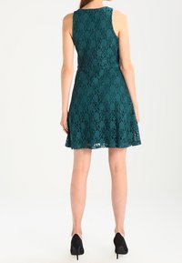 Tropfblaues Spitzenkleid mit ärmellosem Design, taillierter Passform und schwingendem Rock. Der Rücken zeigt filigrane Spitzenverzierungen und endet knapp über dem Knie.
