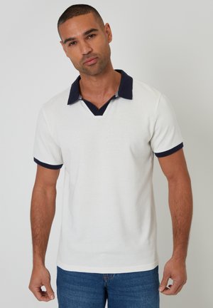 Mand med kort hår, iført en hvid kortærmet polo med navy krave og ærmekanter, kombineret med blå jeans.