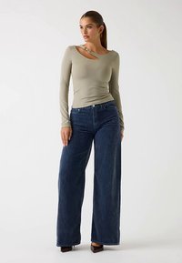 Olivengrøn langærmet top med udskæring foran og dekorative beslag, kombineret med højtaljede, vide blå denimjeans.