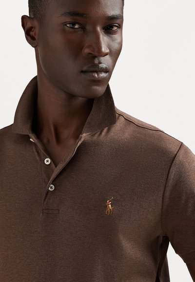 Camisa polo castanha com colarinho, apresentando três botões e um pequeno logótipo bordado do lado esquerdo. Textura suave com ligeiro brilho.