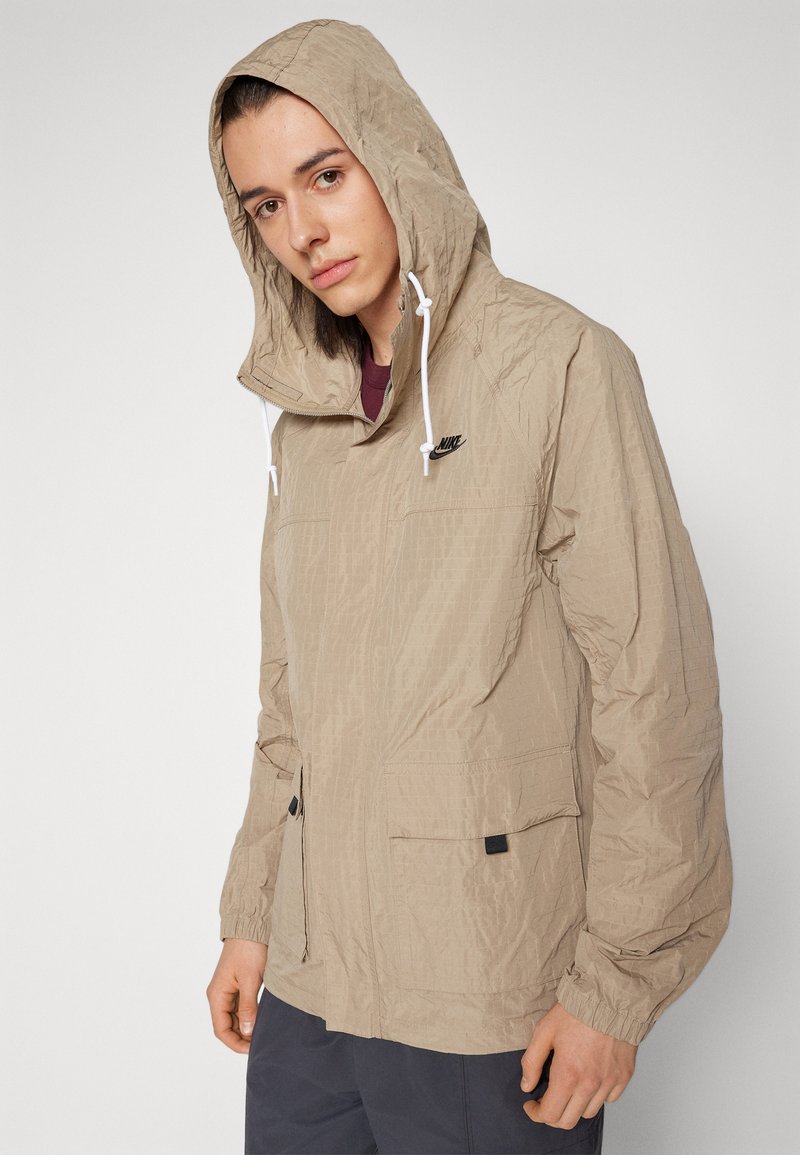 Chaqueta ligera de nylon beige con capucha, bolsillos frontales, puños elásticos y un pequeño logo negro en el pecho. Superficie texturizada.