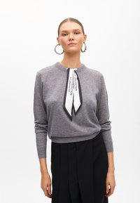 Szary sweter z miękkiego materiału, z okrągłym dekoltem, z czarno-białym detalem w postaci wstążki przy kołnierzu oraz długimi rękawami.