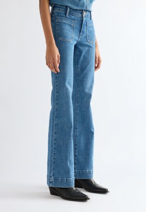 Personne portant un jean bleu taille haute droit et des bottines noires à bout pointu, debout devant un fond blanc uni.