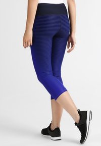 Fioletowe legginsy capri z poziomymi paskami, szerokim czarnym pasem w talii i gładką, elastyczną strukturą. Noszone z czarnymi butami sportowymi.