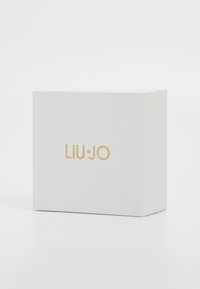 Weißer, strukturierter rechteckiger Kasten mit goldenen "LIU·JO"-Logo, das zentriert auf der Front vor einem schlichten weißen Hintergrund steht.
