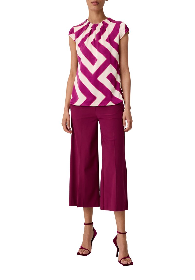 Top a maniche corte in un motivo zigzag rosa e bianco, abbinato a pantaloni larghi color bordeaux scuro e scarpe con tacco aperte coordinate. Tessuto morbido.