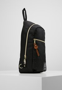 Zaino in tessuto nero con design curvo, zip dorata e dettaglio in pelle marrone. Caratterizzato da una tasca laterale e da una maniglia di trasporto superiore.