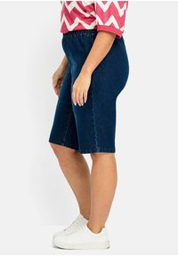 Tmavě modré bermudy z denimu s elastickým pasem, sahající po kolena, s rovým střihem a bezšvovým designem. Kombinovány s bílými teniskami.