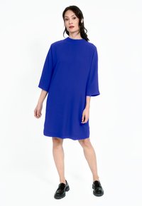 Lila, överdimensionerad klänning med hög hals och ärmar i trekvarts längd. Mjukt tyg, knälång fåll, kombinerad med svarta robusta skor.