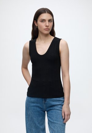 Femme aux cheveux lisses et foncés portant un top noir sans manches et un jean bleu, debout avec une main derrière le dos sur un fond uni.
