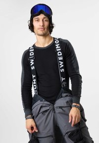 Uomo con camicia a maniche lunghe nera e grigia, pantaloni da neve grigi con bretelle e occhiali da sci sulla fronte, su sfondo semplice.