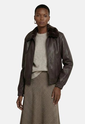 Femme portant une veste en cuir marron foncé avec col en fourrure, un pull beige et une jupe marron texturée, debout devant un fond uni.