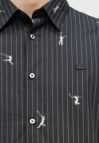 Chemise noire à rayures avec broderie blanche d'un joueur de tennis, boutons contrastants et petit logo Lacoste sur la poitrine.