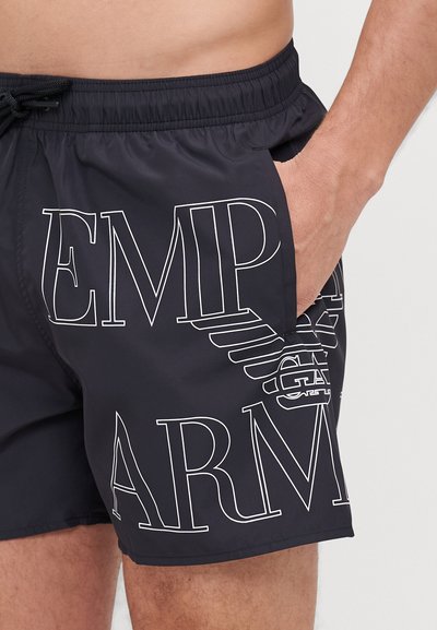 Shorts de bain noirs avec une taille élastique, présentant un grand texte blanc contrastant et un logo sur le côté. Comprend un détail de poche latérale.
