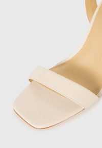 Beige sandals with a square toe, smooth matte finish, single strap across the foot, and a subtle stitched detail along the edges.