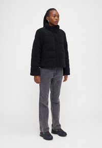 Svart fleece pufferjakke med høy krage, med glidelås og horisontal søm; kombinert med grå jeans og svarte sneakers.