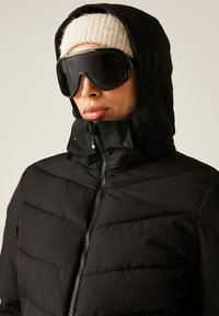 Persoon met een zwarte winterjas met capuchon, een witte gebreide beanie en grote zwarte skibril die recht vooruit kijkt.