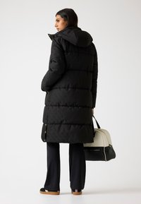 Svart puffjacka med huva, med quiltad textur och guldknappar. Buren över mörka byxor, tillsammans med en handväska i två färger.