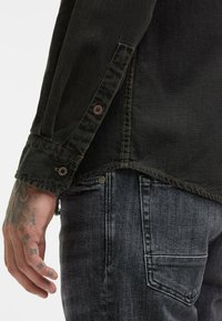 Tatowierte Hand neben dem Knopf eines schwarzen Denim-Hemds, kombiniert mit einer Tasche und Nähdetails von verwaschenen schwarzen Jeans.