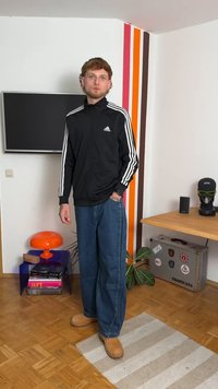 Casaco de treino preto com fecho éclair e riscas brancas, jeans de ganga soltos e botas de camurça castanhas. Em pé sobre um tapete riscado com uma decoração colorida ao fundo.