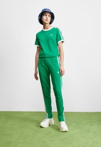 Green athletic tracksuit with white accents and side stripes, made of smooth fabric. Paired with white chunky sneakers and a blue bucket hat.
