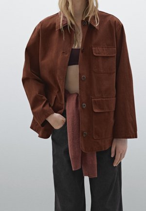 Person wearing a brown buttoned jacket over a cropped top, with a maroon sweater tied at the waist and black pants.