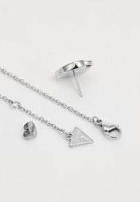 Silver earrings featuring a stud and a chain with a triangular pendant and clasp. Smooth texture with engraved details on the pendant.