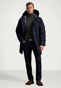 Mężczyzna w granatowym parku z kapturem, ciemnozielonym swetrze z golfem, ciemnych dżinsach i brązowych mokasynach, stojący z jedną ręką w kieszeni.