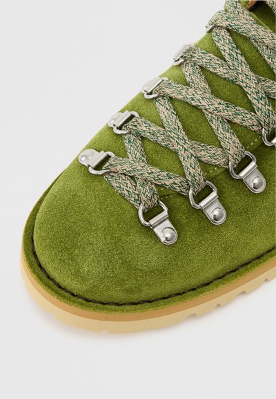 Vihreät nubuk-nahka vaelluskengät, joissa on metalliset nauhakohdat ja kudotut nauhat, varustettuna kumipohjalla ja kontrastivärisellä vaaleanruskealla ulkopohjalla.