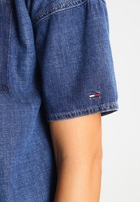 Denim-paita lyhyillä hihoilla, syvänsininen väri, vaaleat ompeleet ja pieni logo-merkki punaisella, valkoisella ja sinisellä hihassa.