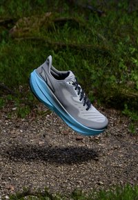 Chaussure de course pour homme avec une tige grise, une semelle intermédiaire bleue et des lacets noirs. Présente une surface texturée et des accents réfléchissants. Design léger.