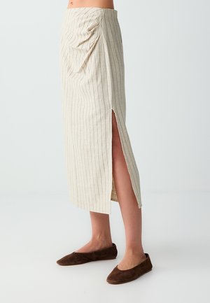 Beżowa spódnica midi w cienkie prążki z bocznym rozcięciem, noszona z brązowymi płaskimi butami, pokazana od talii do stóp na jasnym tle.