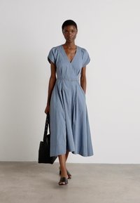 Lichtblauwe jurk met korte, afgekorte mouwen, V-hals, cinched taille en vloeiende rok. Gecombineerd met een zwarte handtas en schuifsandalen.