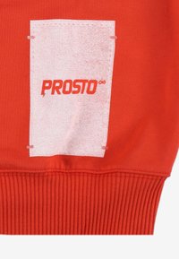 Czerwona bluza z kapturem z białą prostokątną łatką z napisem "PROSTO" w grubym czerwonym kolorze. Mankiety i dół bluzy są prążkowane, co dodaje faktury.