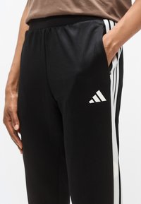 Μαύρο αθλητικό παντελόνι με ελαστική μέση, διακοσμημένο με λευκές ρίγες στο πλάι και ένα μικρό λευκό λογότυπο στο μπούτι. Λείο, ελαστικό ύφασμα.