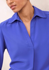 Bluza z V-izrezom v živahni modri tkanini s gladko teksturo in standardnim ovratnikom. Ponuja strukturirano prileganje in subtilen šivni detajl.