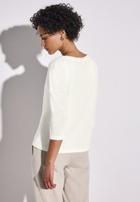 Blusa bianca strutturata con maniche a tre quarti, design pullover e scollatura rotonda, abbinata a pantaloni beige. Tessuto morbido.