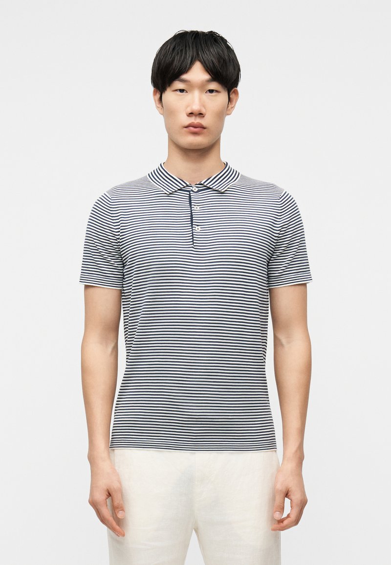 Jeune homme debout, portant un polo à manches courtes à rayures horizontales bleu marine et blanches, pantalon de couleur claire, fond neutre.