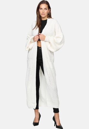 Cardigan lungo bianco lavorato a maglia con maniche a palloncino ampie, due tasche frontali, design aperto e motivo testurizzato. Abbinato a un abbigliamento nero.