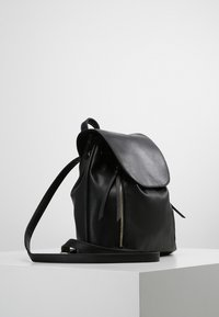 Sac à dos en cuir noir avec un rabat supérieur arrondi, un détail de fermeture éclair latérale et une bretelle élégante. Texture lisse et design moderne.