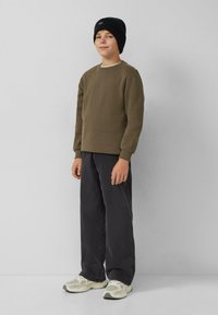 Chłopak stoi w czapce beanie w kolorze czarnym, oliwkowej bluzie, luźnych czarnych spodniach i białych sneakersach na tle jednolitego jasnoszarego tła.