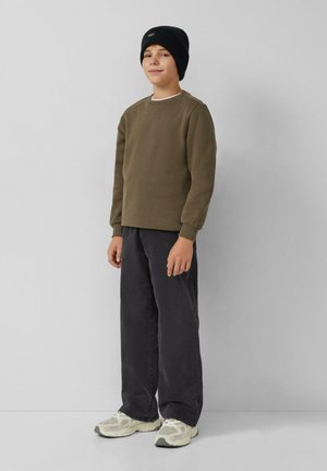 Junge steht da und trägt eine schwarze Mütze, einen olivgrünen Sweatshirt, lockere schwarze Hosen und weiße Sneaker vor einem schlichten hellgrauen Hintergrund.