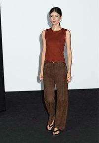 Bruine mouwloze top en bijpassende brede suède broek. Model draagt zwarte slippers; minimalistische setting met neutrale achtergrond.