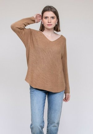 Jeune femme aux cheveux bruns raides portant un pull marron ample et un jean bleu, posant avec une main près de la tête.