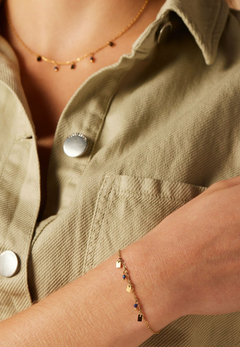 Gouden ketting en armband met kleine geometrische bedels en blauwe accenten, tegen een gestructureerd, olijfgroen shirt.