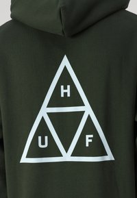 Sudadera verde oscura con un gran gráfico triangular blanco en la parte posterior que presenta las letras H, U y F dispuestas dentro de los triángulos.
